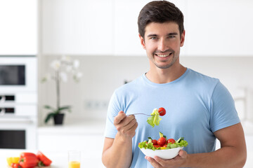 Glad unshaven smiling man in the kitchen holds a plate with a vegetable salad in his hands, looks good wearing a blue T-shirt. Healthy and vegetarian food