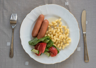 Boiled sausages with pasta and vegetable salad on a plate on the table.
