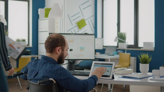 Paralyzed manager, businessman with disabilites handicapped immobilized man using computer and tablet in same time working in business start-up office sitting in wheelchair. Busy multitasking employee