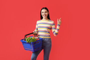 Young woman with shopping basket pointing at something on color background
