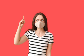 Young woman wearing medical mask and pointing at something on color background