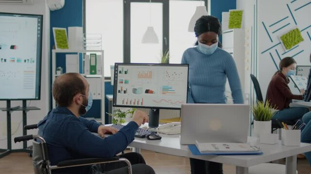African manager with face mask working with handicapped invalid immobilized colleague in new normal business office during covid19 global pandemic, calculating profit on laptop taking notes on