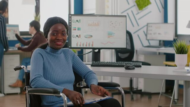 Happy african businesswoman with disabilities looking at camera smiling in immobilized paralysed in wheelchair in business economic office, working on pc with data prosessing financial information