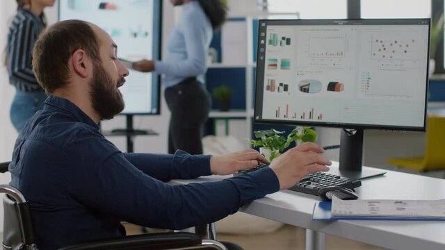 Handicapped disabled businessman in wheelchair discussing with colleague, asking informations typing on computer, checking statistic datas working in start up business office with diverse team