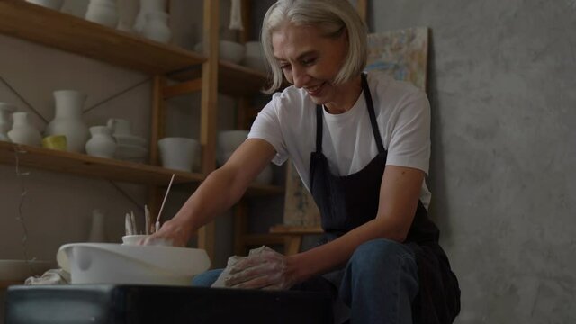 Aged female artisan spending daytime leisure for creating manufacturing handmade, skilled sculptor enjoying clay modeling and pottery hobby in art studio