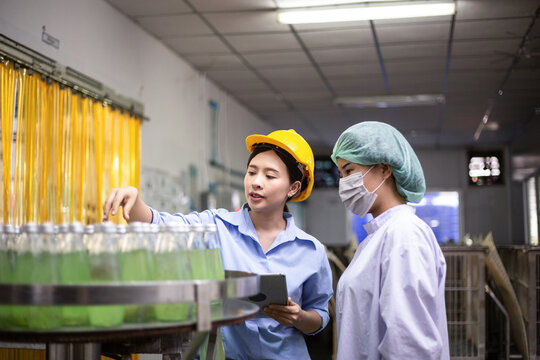 Asian female manager and worker using digital tablet checking information and quality control for juice drink product on bottling factory machinery. Process of beverage industrial production
