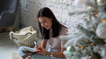 Happy Asian female with textbook writing wish list to Santa Claus during holidays time in home interior, cheerful hipster girl with diary making notes of dreams for ask St Nicolas enjoying Christmas