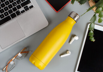 Yellow insulated bottle on grey desk surrounded by modern gadgets and plant