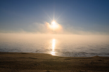 Star like sun with rays of light behind fog on sea at sunset with calm water and beach
