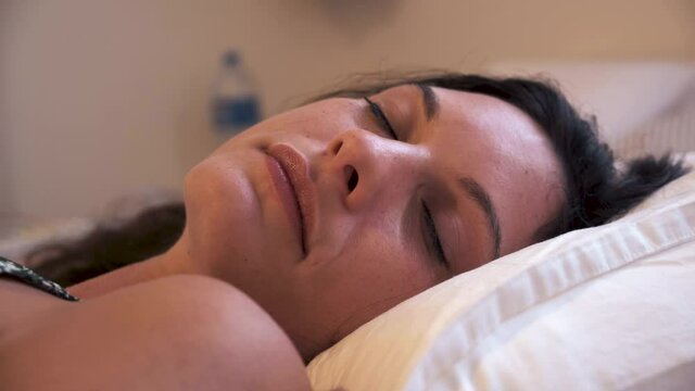 woman falling sleeping in bed. head on a pillow. Young beautiful lady laying relaxed with her eyes opened and then closed. siesta time sensual sexy girl taking a nap. noon summertime white beddings.