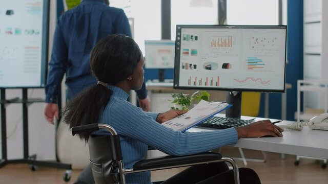 African woman with disabilities immobilized in wheelchair working on computer checking reports, analysing datas from clipboard. Multiethnic team discussing in strat up business financial office