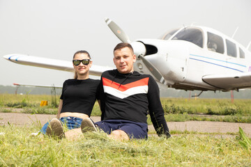 young man and woman sit on the grass on the background of an old small plane at the airfield in Ukraine in the city of Dnipro