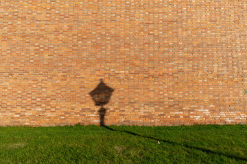 the shadow of a street lamp against a green lawn and a brick wall at sunset