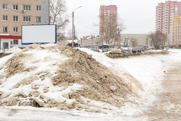A large pile of dirty snow on the side of the road with passing cars. In the background are high-rise buildings of a modern metropolis. Against the background of a gray cloudy sky.