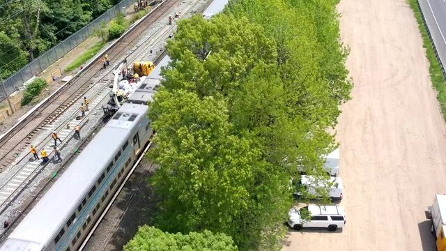 an aerial time lapse over men repairing train tracks on a sunny day. The drone camera focuses on a yellow truck on the tracks and orbits to the left above the trees as people work on the tracks.