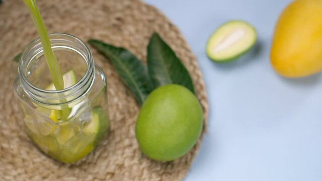 Refreshing raw mango juice / syrup with sliced lemon pouring in a bottle - Aam Panna  Summer drink in India. Closeup shot of a ripe and a raw mango kept together on a jute mat on a wooden table - c...