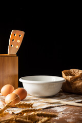 Top view of pastry objects, white bowl, eggs, flour and ears of wheat on cloth and wooden table, selective focus, vertical