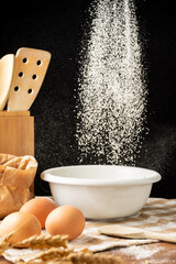 Fototapeta na wymiar View of flour falling on white bowl with rustic wooden objects, on wooden table, on black background, in vertical