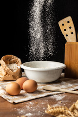 View of flour falling on white bowl, on wooden table with eggs, wooden spoons, selective focus, on black background, vertical