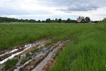 Rural muddy dirt road in summer after rain on the background of green bushes. Off-road, you can see the village in the distance