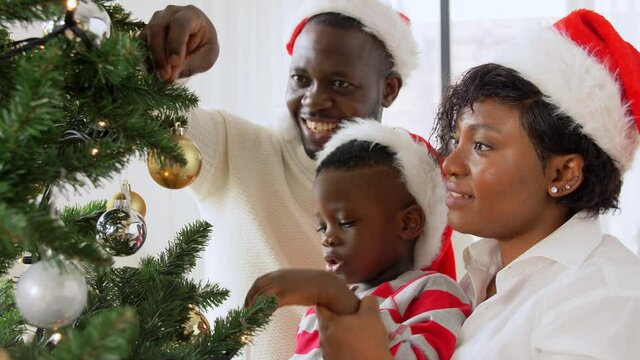 family, winter holidays and people concept - happy african american mother, father and baby son decorating christmas tree at home on