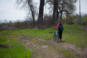 Fototapeta premium a girl with a bicycle walks a field trail. Woman riding a mountain bike in the forest. Hiker woman with backpack. one young woman riding a bike in nature. rolling the bike in his hands. back view.