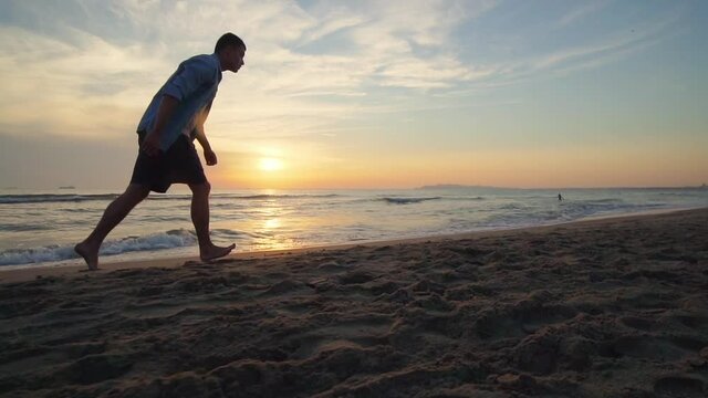 A male athlete is making back flip and lands on feet on the sandy tropical beach at sunset, slow motion