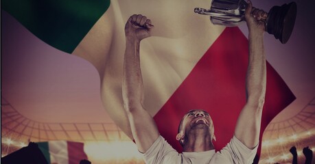 Male athlete with arms in air holding cup at sports stadium over italian flag