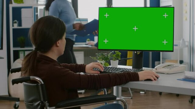Disabled handicapped immobilized woman emplyee working on desktop computer with chroma key display, green mock-up screen, greenscreen pc, typing project informations sitting in wheelchair.