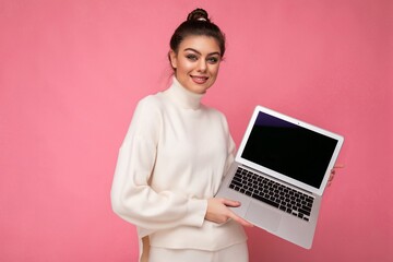 Photo of beautiful smiling brunet young woman with gathered dark hair wearing white sweater holding computer laptop and looking at camera isolated on pink wall background