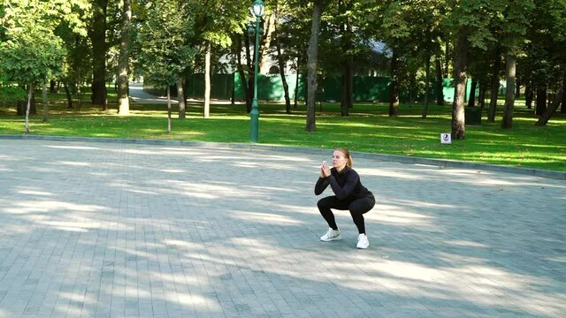 Slow motion fit woman wearing black sportswear jumping and squatting on paved court on sunny day. Arc shot female athlete working out in park. Concept of sport