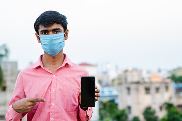 Young Indian man wearing surgical mask and showing smartphone to camera.