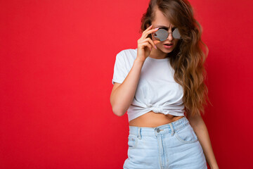Fototapeta premium Shot of charming sexy young dark blonde curly woman isolated over red background wall wearing casual white t-shirt and stylish sunglasses looking at camera