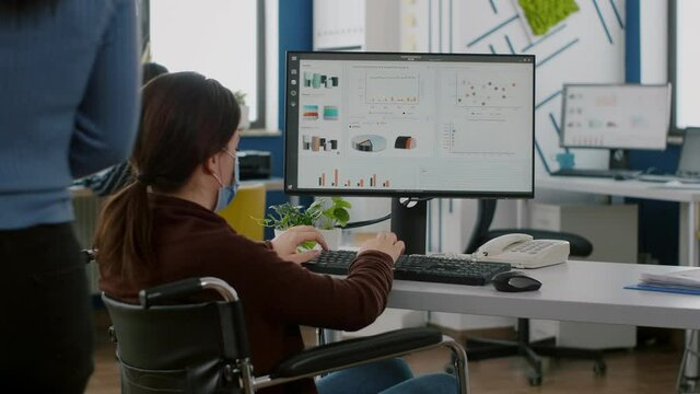 Disabled handicapped businesswoman with face mask immobilized in wheelchair working at financial project typing on pc showing data, prosessing on display sitting in new normal startup business office.
