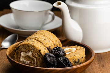 Side view of homemade prune sponge cake roll and dried pitted prune fruits in wooden bowl with white ceramic coffee pot, a cup of coffee and metal spoon on wooden table.