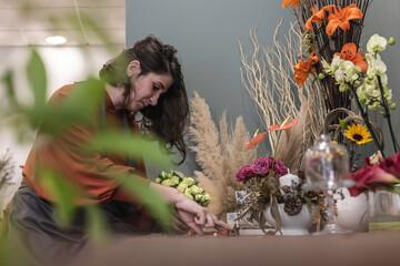 View of a young woman working as florist in her flower shop, sitting at wooden table and creating flower decoration, businesswoman concept. - obrazy, fototapety, plakaty