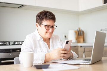 Obraz na płótnie Canvas Happy senior woman using mobile phone while working at home with laptop. Smiling cool old woman wearing eyeglasses messaging with smartphone.