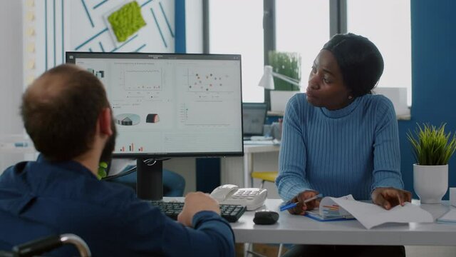 Handicapped businessman and african employee working at financial project discussing in start up company, checking statistic info looking at pc showing data prosessing on display for regarding economy