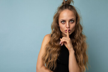 Photo of young beautiful blonde curly woman with sincere emotions wearing black top isolated over blue background with copy space and showing shhh gesture. Be quiet