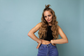 Young cool sexy beautiful winsome blonde curly woman with sincere emotions wearing stylish black top isolated over blue background with copy space