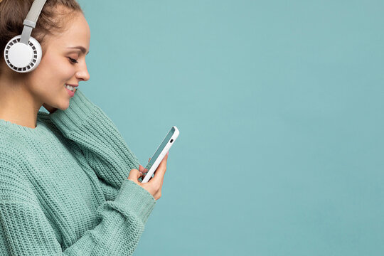 Closeup Photo Of Beautiful Joyful Smiling Young Woman Wearing Stylish Casual Clothes Isolated Over Background Wall Holding And Using Mobile Phone Wearing White Bluetooth Headphones Listening To Music