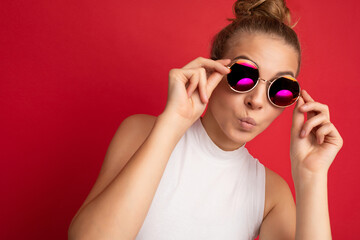 Photo shot of beautiful young blonde woman wearing casual clothes and stylish sunglasses isolated over colorful background looking at camera and giving kiss