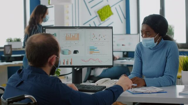 Multiethnic businessteam with face masks working in new normal startup company office during coronavirus epidemic, checking business reports, disabled leader sitting in wheelchair briefing ideas