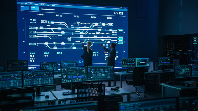Project Manager and Computer Science Engineer Talking while Using Big Screen Display Showing Infrastructure Infographics and Data.Telecommunications Company System Control and Monitoring Room