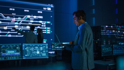 Handsome Confident Male Project Manager works on a Laptop Standing in System Control Room. In...