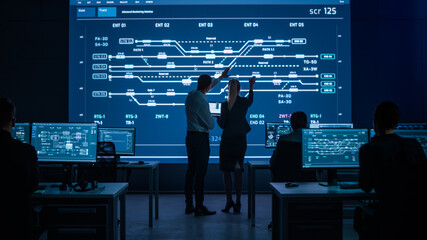 Project Leader and Chief Executive Discuss Data Shown on Screen. In Background Big Screen Showing Infographics of Infrastructure, Charts, System Analysis. Control Room Professionals Working