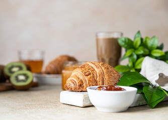 Breakfast with a cup of coffee, fresh crispy croissant, jam, kiwi and juice, light stone background. Summer breakfast.