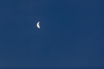 An incomplete disc of the moon in the form of a white crescent against a dark blue cloudless sky
