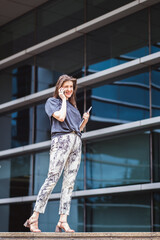 Beautiful woman with brown long hair in elegant casual clothes with a laptop speaks on a mobile phone outdoors with office buildings in the background