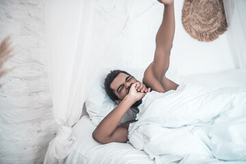 Obraz na płótnie Canvas Darkskinned yawning man lying in white bed